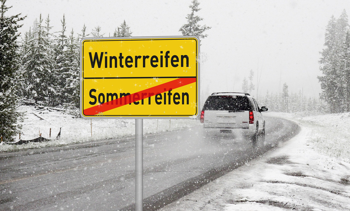 Vehicle on a snow-covered road, in front of which a place-name sign on the summer tire is crossed out and winter tires can be read